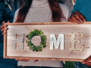 Two people holding a rustic wooden HOME sign with a wreath, representing first-time homebuying in the Triad region of North Carolina